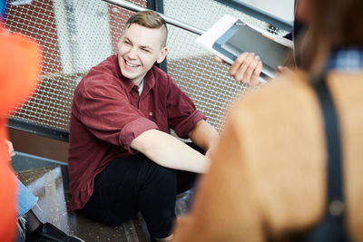 Smiling man sitting on steps while talking to friends in university