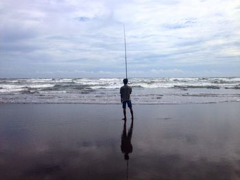 Rear view of man with fishing rod standing on shore at beach against cloudy sky