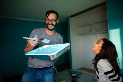 Happy father holding paint tray and brush while daughter laughing at home