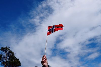 Low angle view of hand holding norwegian flag against sky