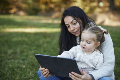 Mother and daughter sitting in autumn park and using digital tablet