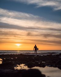 Man with surfboard standing on shore at beach during sunset