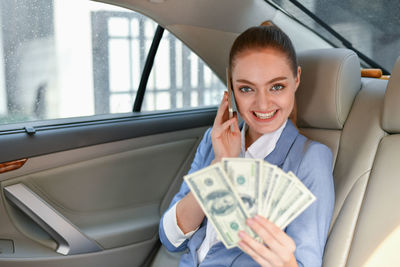 Portrait of smiling businesswoman with paper currency talking on phone while sitting in car