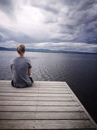 Rear view of woman sitting on jetty over sea against cloudy sky