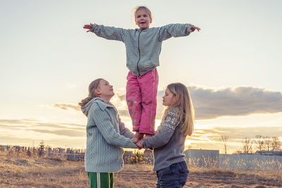 Girl standing on sisters hands against sky during sunset