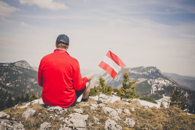 Rear view of man holding flag while sitting on mountain against sky