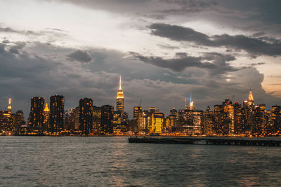 Illuminated buildings by river against cloudy sky