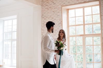 Groom holding flower bouquet with bride standing by window at home