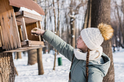 A teenager pours seeds into a homemade bird feeder on a tree in a winter park. animal care.