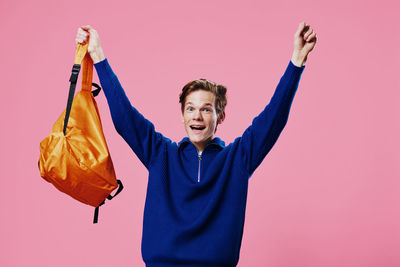 Portrait of smiling young man holding backpack standing against pink background