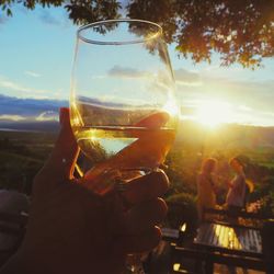 Close-up of hand holding beer glass against sunset