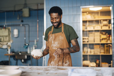 Smiling male baker dusting flour on table and holding dough while working at bakery
