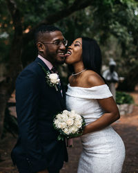 Bride licking groom cheek against trees