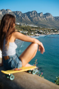 Rear view of woman sitting by sea against mountains