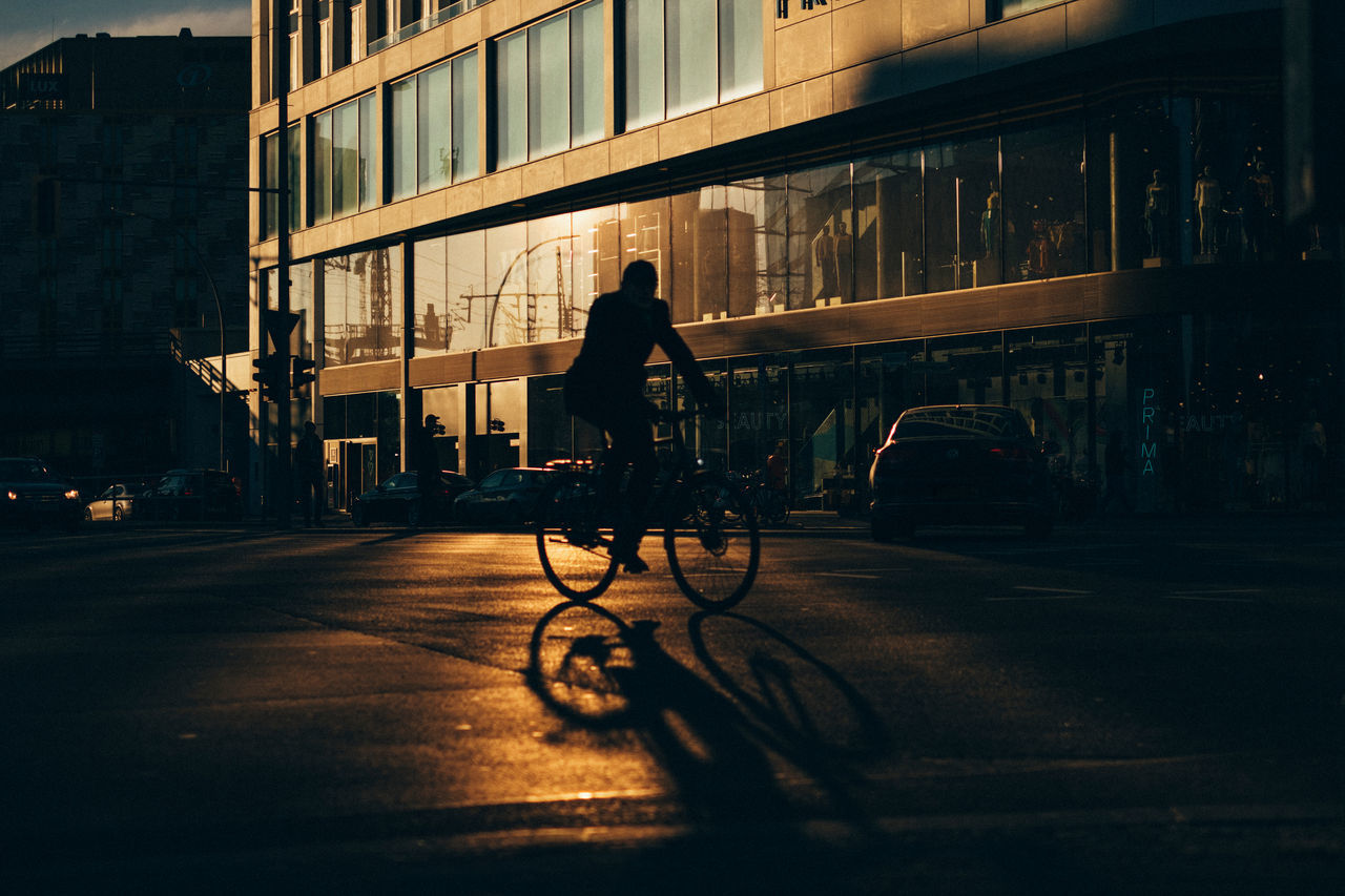 city, architecture, building exterior, transportation, built structure, mode of transportation, street, bicycle, one person, real people, city life, building, land vehicle, full length, motor vehicle, car, walking, sunlight, riding, place