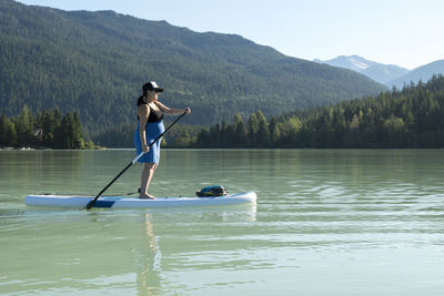 Side view of barefoot woman riding sup board on lake against green mountain ridge on sunny summer day in british columbia, canada