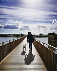 Rear view of woman with dog walking on footbridge over river against sky