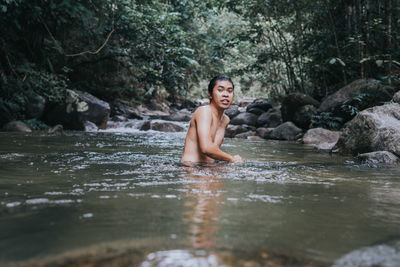 Portrait of shirtless man sitting on rock in forest