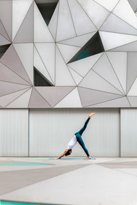 Full body adult man in sportswear performing three legged downward facing dog exercise while doing yoga against wall with geometric ornament