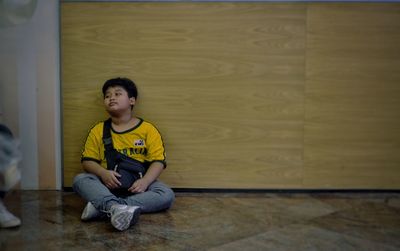 Boy looking away while sitting on floor against wall