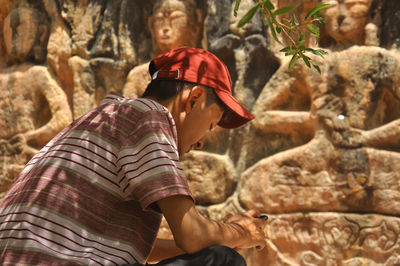 Rear view of a guy using phone while sitting in front of gyalwa ringna 5 dhyani buddha rock statue