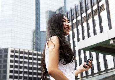 Smiling young woman holding mobile phone while standing in city