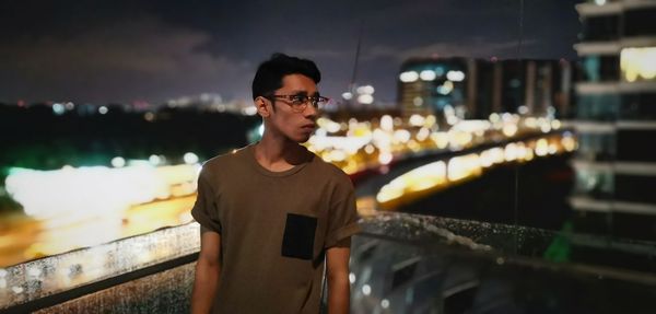Thoughtful young man looking away while standing against illuminated city at night