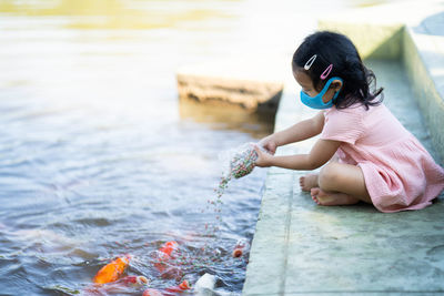 Cute girl wearing protective face mask feeding carp koi fishes at pond.