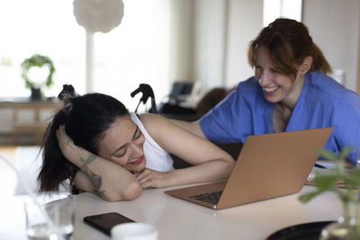 Happy female caregiver talking to woman with paraplegia watching laptop at home