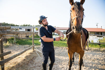 Adult male in protective helmet holding stallion by reins against stables of riding school in countryside