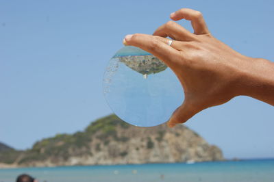 Cropped image of hand holding crystal ball with reflection against blue sky