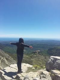 Rear view of woman standing with arms outstretched on rock against clear sky