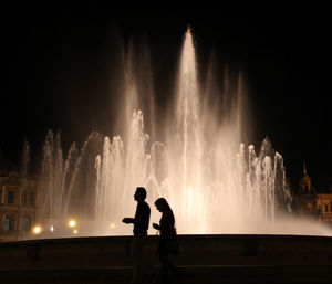 Silhouette friends walking by fountain in city against clear sky at night