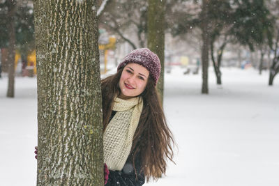 Portrait of young woman standing behind tree trunk during winter