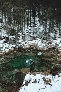 Man standing on rock by pond in forest during winter