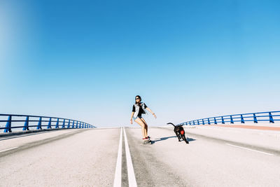 Dog running with woman skateboarding against blue sky