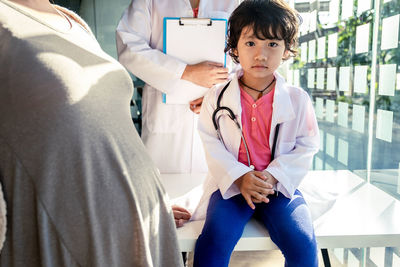 Portrait of cute girl sitting on table while doctor and pregnant woman standing in clinic