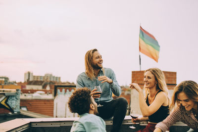 Cheerful friends talking while enjoying wine together on terrace during party against sky