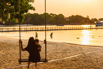 Rear view of couple sitting on swing at beach during sunset