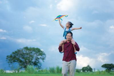 Father giving piggy back ride to daughter holding kite against cloudy sky