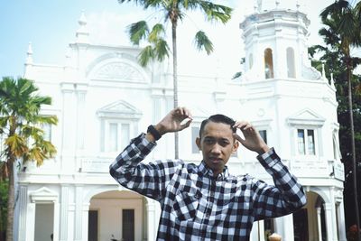 Portrait of young man standing by palm tree and building