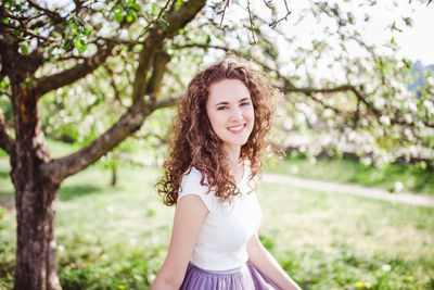 Happy curly hair young woman under the blooming apple tree. smiling, looking at the camera.