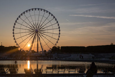 Silhouette ferris wheel by river against sky during sunset