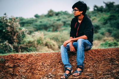 Young man looking away while sitting on field against trees