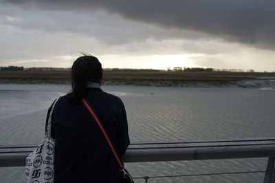 Rear view of woman standing on pier at beach against cloudy sky