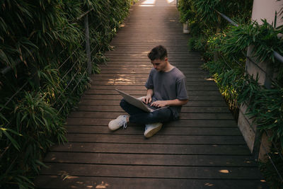 From above of concentrated young male student in stylish outfit typing on laptop while sitting on wooden walkway surrounded by tropical plants during online lessons in sunlight