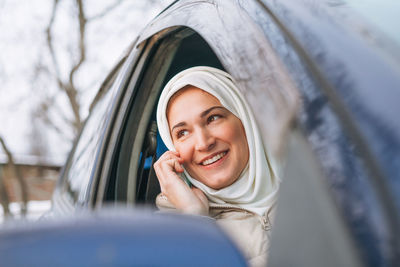 Beautiful smiling young muslim woman in headscarf using mobile in right-hand-drive car