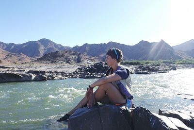 Woman sitting on rock by mountains against clear sky