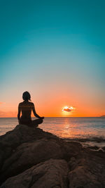 Man sitting on rock at beach against sky during sunset