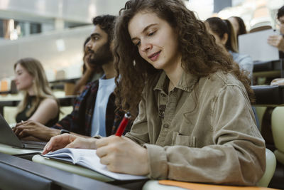 Curly haired young woman writing in book while sitting in classroom at college